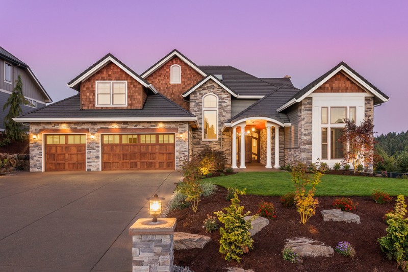 A large, modern suburban house with stone and wood exterior, three-car garage with wooden doors, a well-manicured lawn, landscaped garden, and a driveway at sunset.