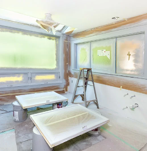 A room undergoing renovation with windows and walls taped and covered for painting. A stepladder and two cabinet doors rest on paint cans, and the floor is covered with protective sheets.