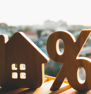 Wooden house and percentage symbol figures are displayed side by side with a blurred cityscape in the background, representing concepts of real estate and interest rates.