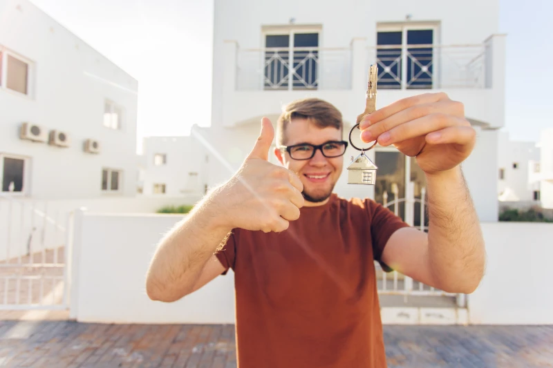 A smiling man wearing glasses holds up a house key and gives a thumbs up, standing in front of a white residential building on a sunny day.